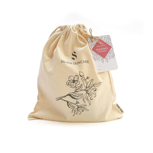 photo of a creamy coloured organic cotton drawstring bag on a white background. the bag has been screen printed with the silvan skincare logo and a botanical drawing of a flower. the bag has a rectangular gift tag on it . the bag can be used to fill with natural skincare gifts or as a toiletry bag for travelling