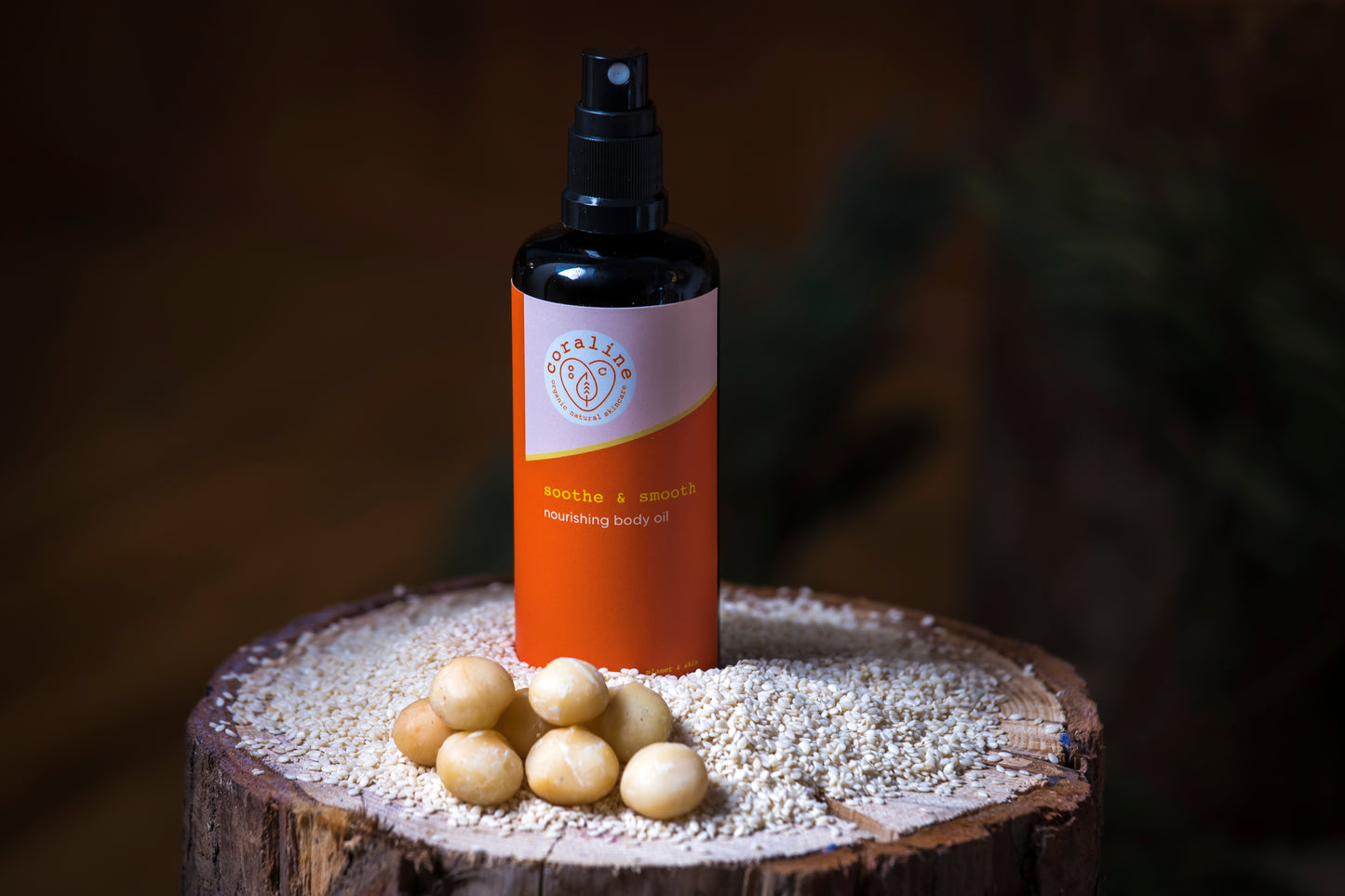 A black bottle of smooth and soothe nourishing body oil, with an orange label, sits on a wooden surface scattered with small, cream-coloured macadamia nuts and grains. The background is softly blurred, giving a warm, earthy ambiance.