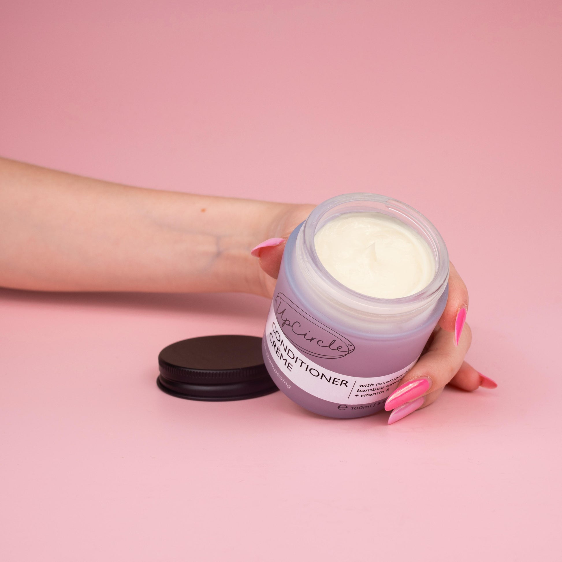 lifestyle photo of upcircle's conditioner cream on a pink background. the conditioner is in a blue/lilac frosted glass jar with black aluminium lid. The jar is open and you can see the creamy texture of the conditioner inside. It is being held by a woman's hand with multi tonal pink finger nails tilted towards the front of the image