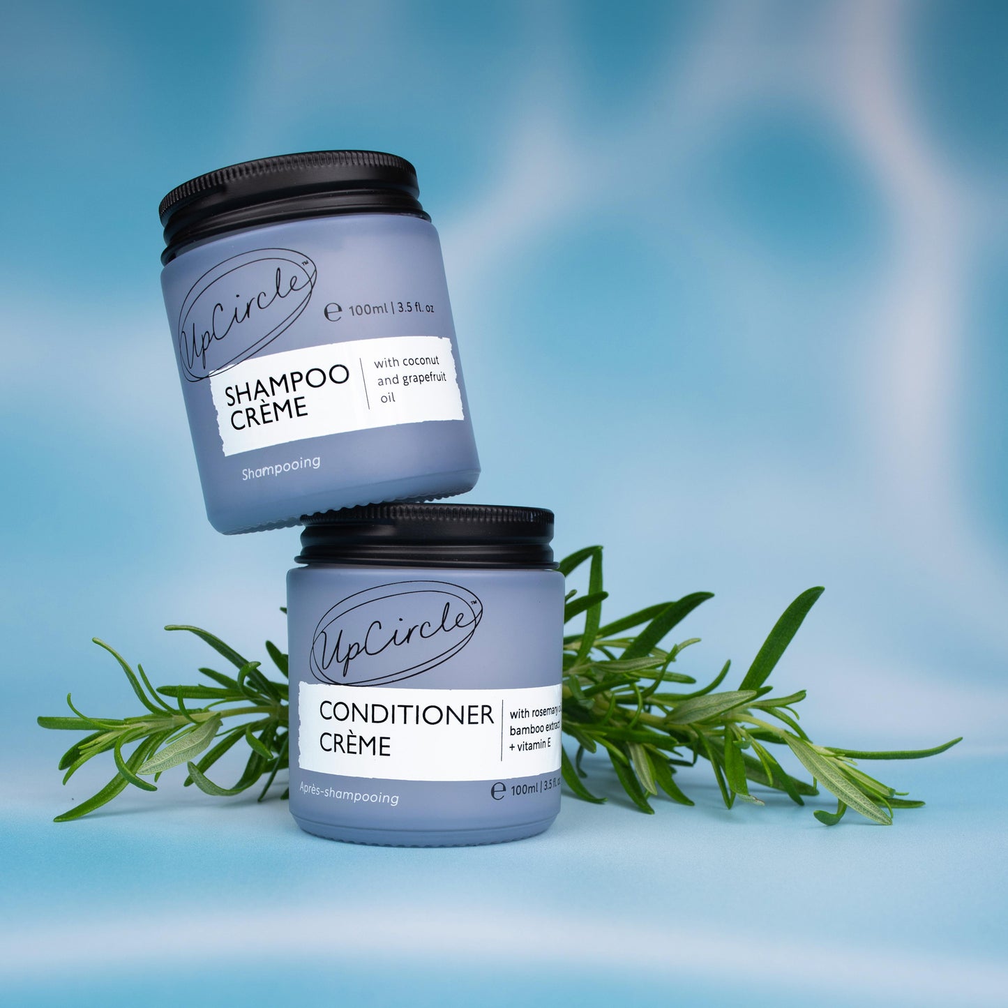 lifestyle photo of upcircle's shampoo creme and conditioner creme on a blue iridescent background. the shampoo is balanced at an angle on top of the conditioner. both products are in light blue glass jars with black aluminium lids. there is a sprig of rosemary in the background