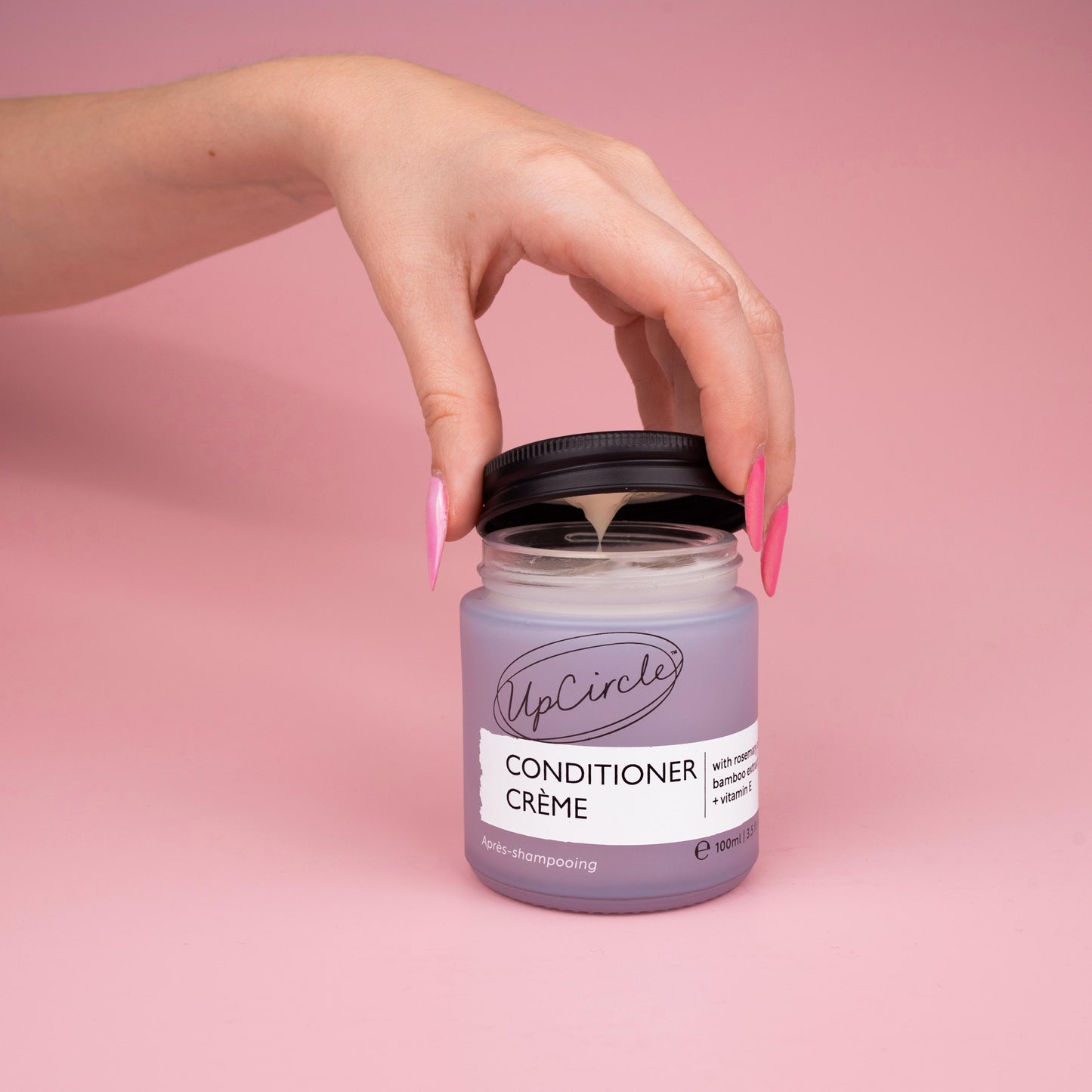 lifestyle photo of upcircle's conditioner cream on a pink background. the conditioner is in a blue/lilac frosted glass jar with black aluminium lid. The jar is open and you can see the creamy texture of the conditioner inside. The lid being lifted off the jar by a woman's hand with multi tonal pink finger nails