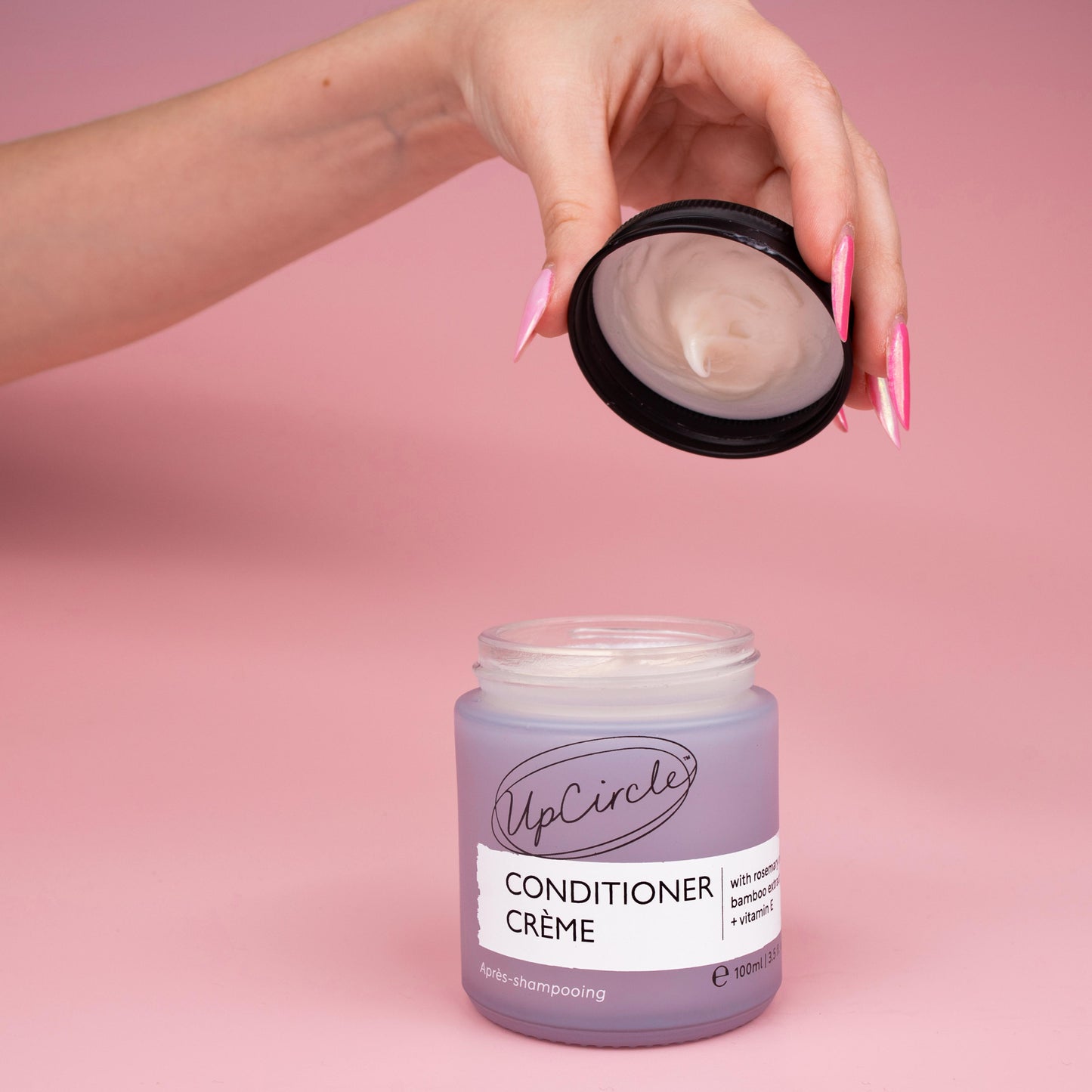 lifestyle photo of upcircle's conditioner cream on a pink background. the conditioner is in a blue/lilac frosted glass jar with black aluminium lid. The jar is open and you can see the creamy texture of the conditioner inside. The lid being lifted off the jar by a woman's hand with multi tonal pink finger nails