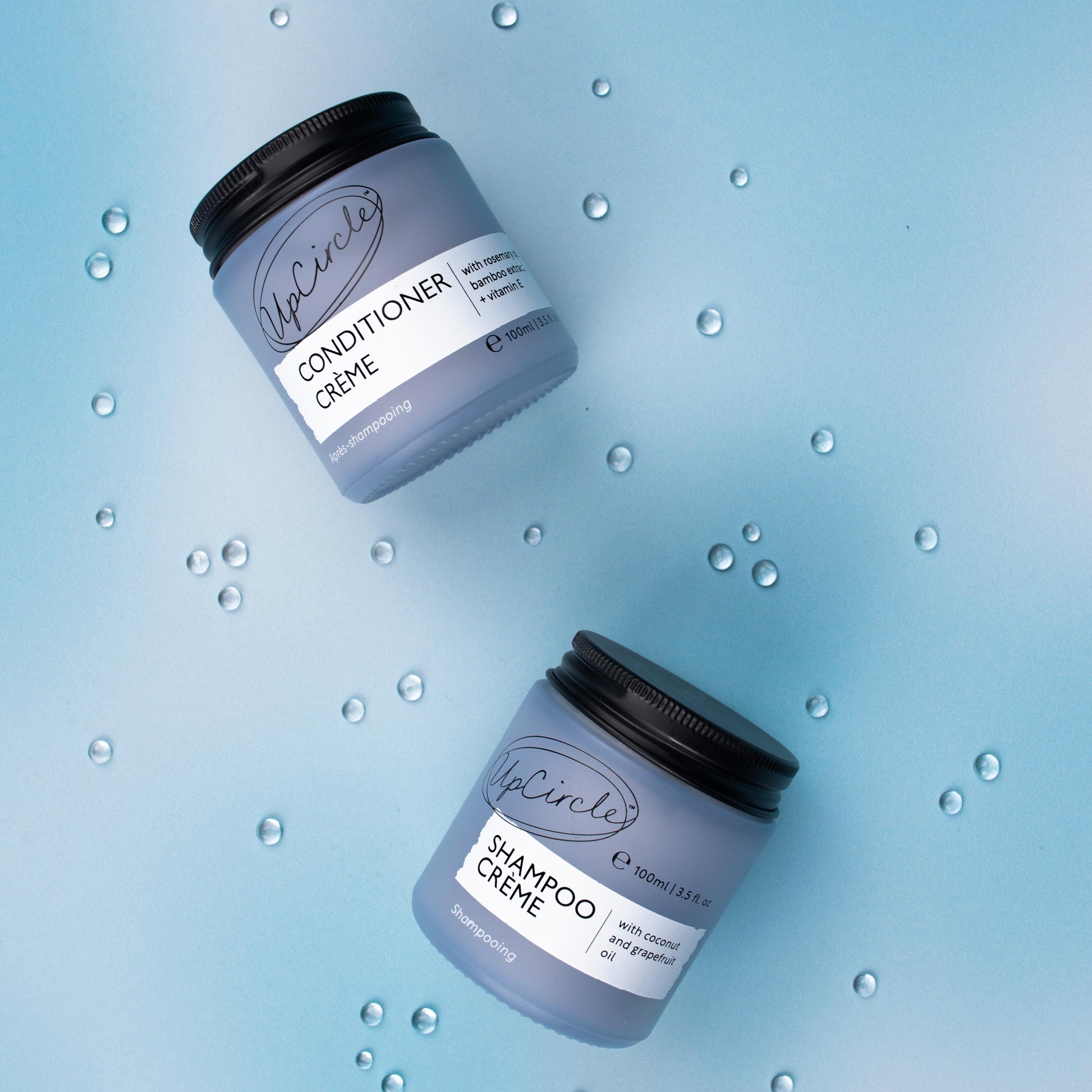 flatlay photo of upcircle's shampoo creme and conditioner creme on a blue iridescent background. both products are in light blue glass jars with black aluminium lids. there are clear water droplets in the background