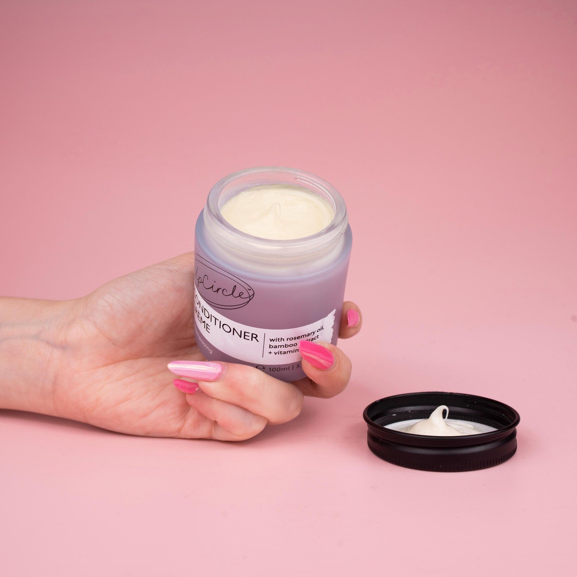 lifestyle photo of upcircle's conditioner cream on a pink background. the conditioner is in a blue/lilac frosted glass jar with black aluminium lid. The jar is open and you can see the creamy texture of the conditioner inside. It is being held by a woman's hand with multi tonal pink finger nails
