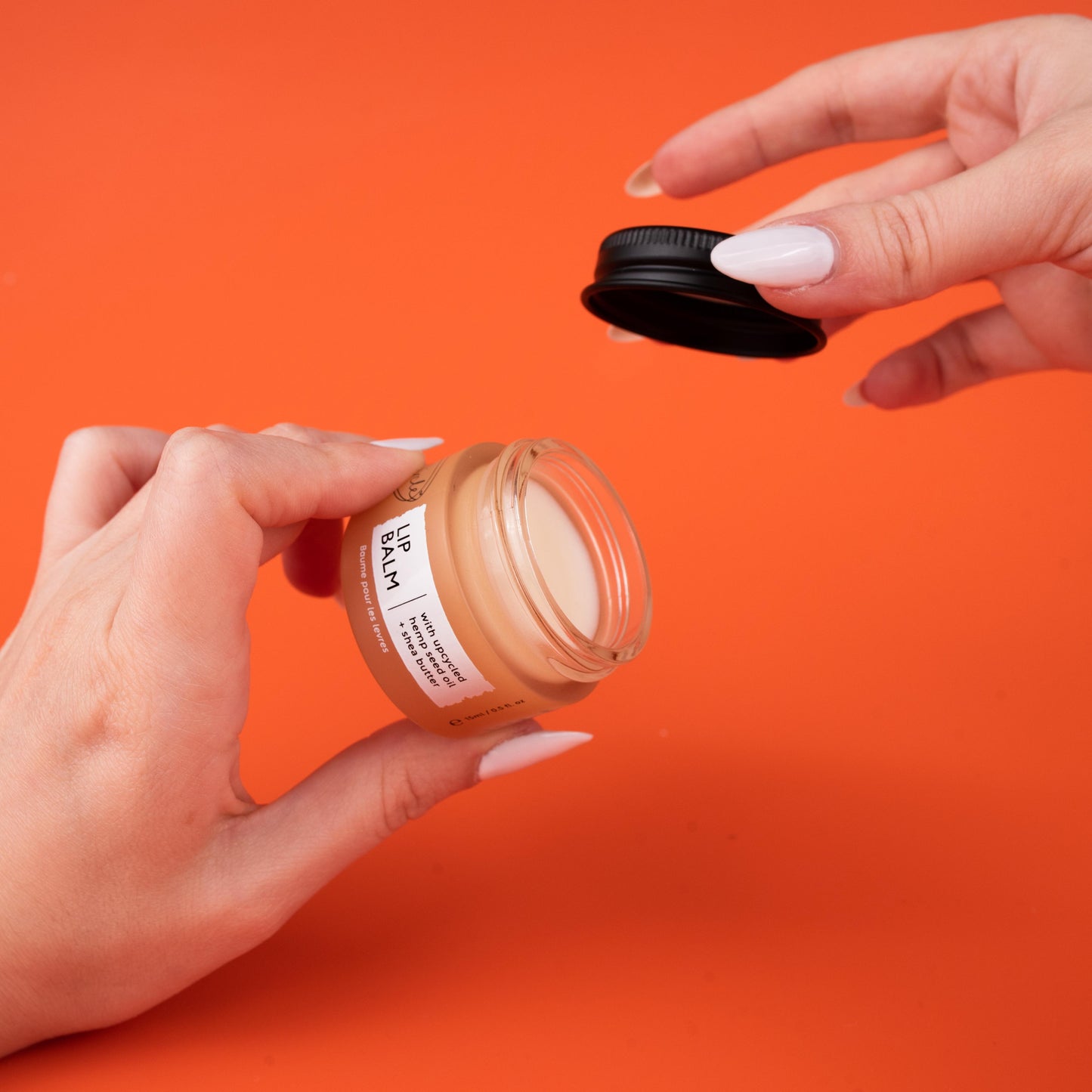 A person's hands are holding an open jar of upcircle lip balm. One hand is holding the jar and the other hand is lifting the black lid. The background is a solid bright orange.
