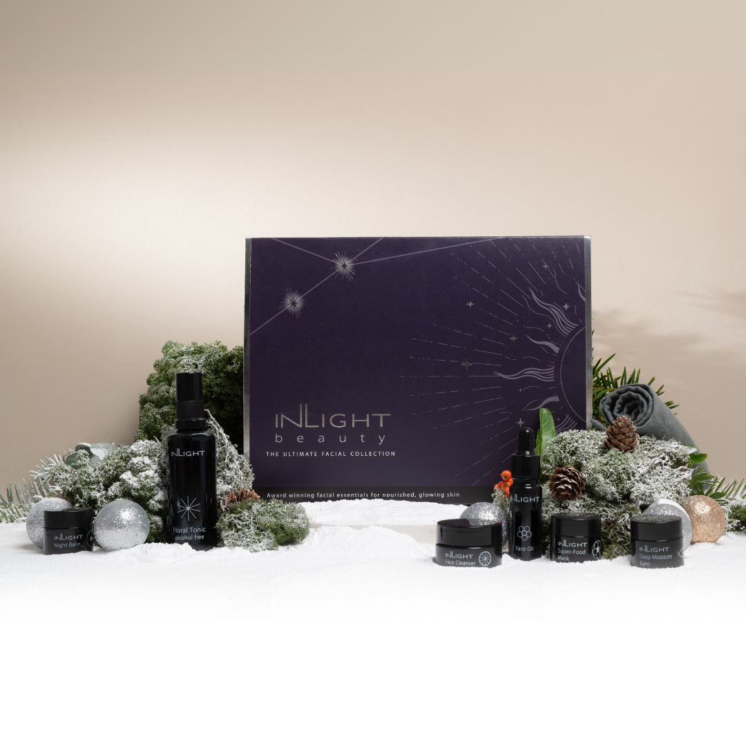festive scene showcasing inlight beauty' ultimate facial collection - award winning facial essentials for nourished, glowing skin. purple and silver box is sitting on white snow with green festive plants pine cones and baubles and the black glass bottles of the product