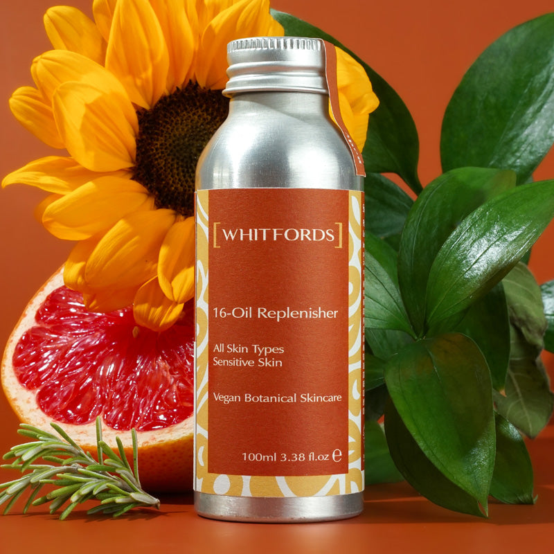 Whitfords skincare body oil in an aluminium bottle with dark orange label on an orange background with dark green leaves, a sunflowers, frond of rosemary and half a grapefruit.. the label reads ' whitfords 16-oil replenisher. all skin types, sensitive skin, vegan botanical skincare'