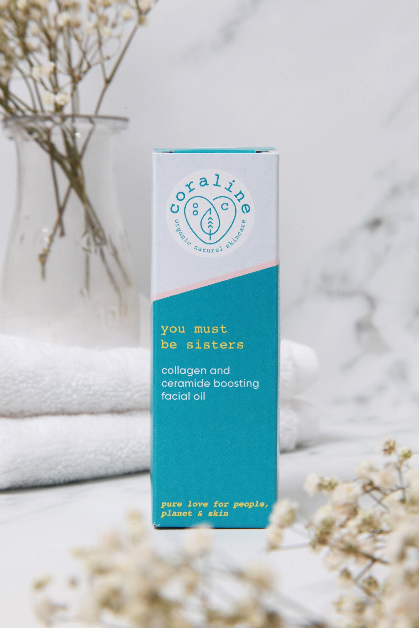 A box of "You Must Be Sisters" facial oil stands in its packaging on a marble surface. Behind them, white towels and delicate baby's breath flowers in a vase. The flowers are blurred out in the foreground too.