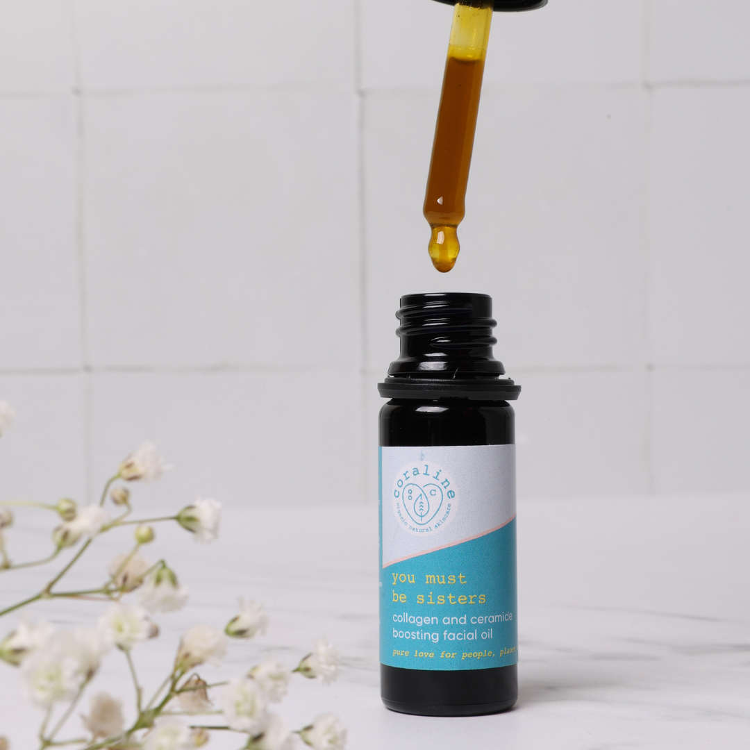 A bottle of "you must be sisters" collagen and ceramide-boosting facial oil is pictured with a dropper dispensing the oil above it. The bottle has a blue and white label. In the foreground, there's a small sprig of white flowers on a marble surface.