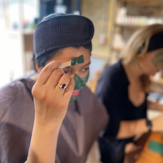 A person is holding a small brush and using it to apply a green face mask to their forehead. They are wearing a black headband and a purple gown. Another person in the background is also wearing the facial mask and a black outfit. 