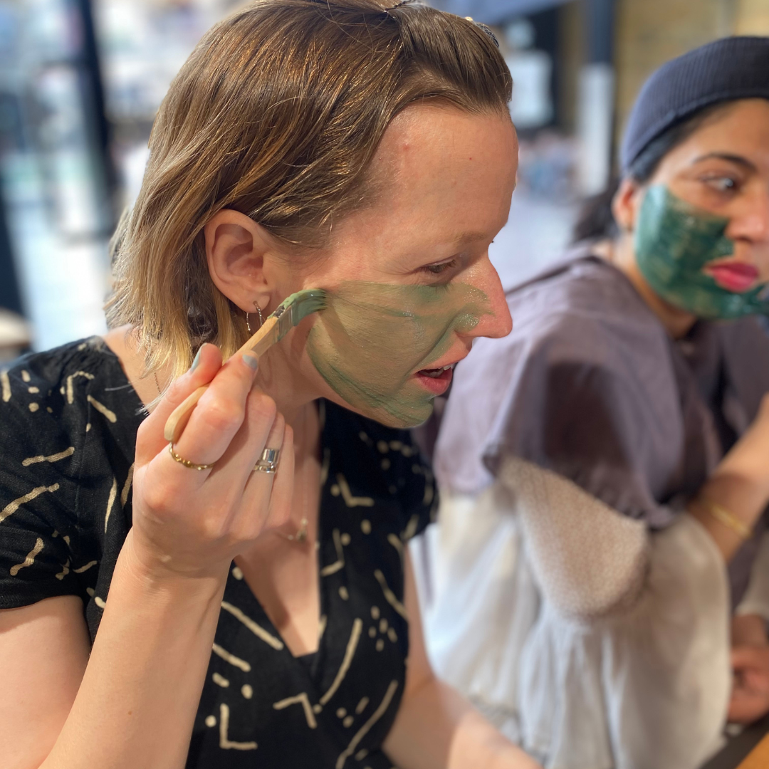 Two ladies shown applying green facial masks with brushes. They are on the Blomma shop floor.