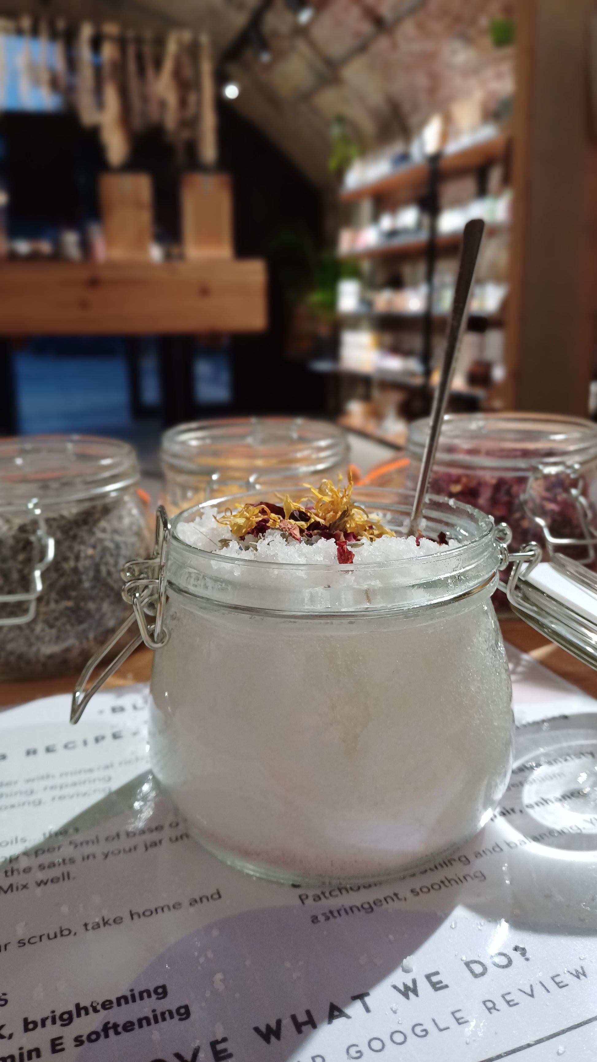 open jar of hand made bath salts with dried flowers. the jars of dried flowers can be seen in the background