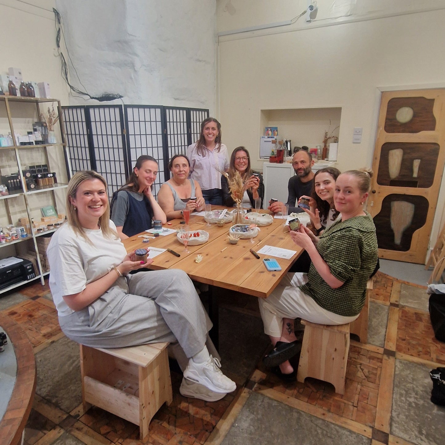 group of people sitting at a wooden table holding up jars of handmade body scrubs after a workshop at blomma beauty
