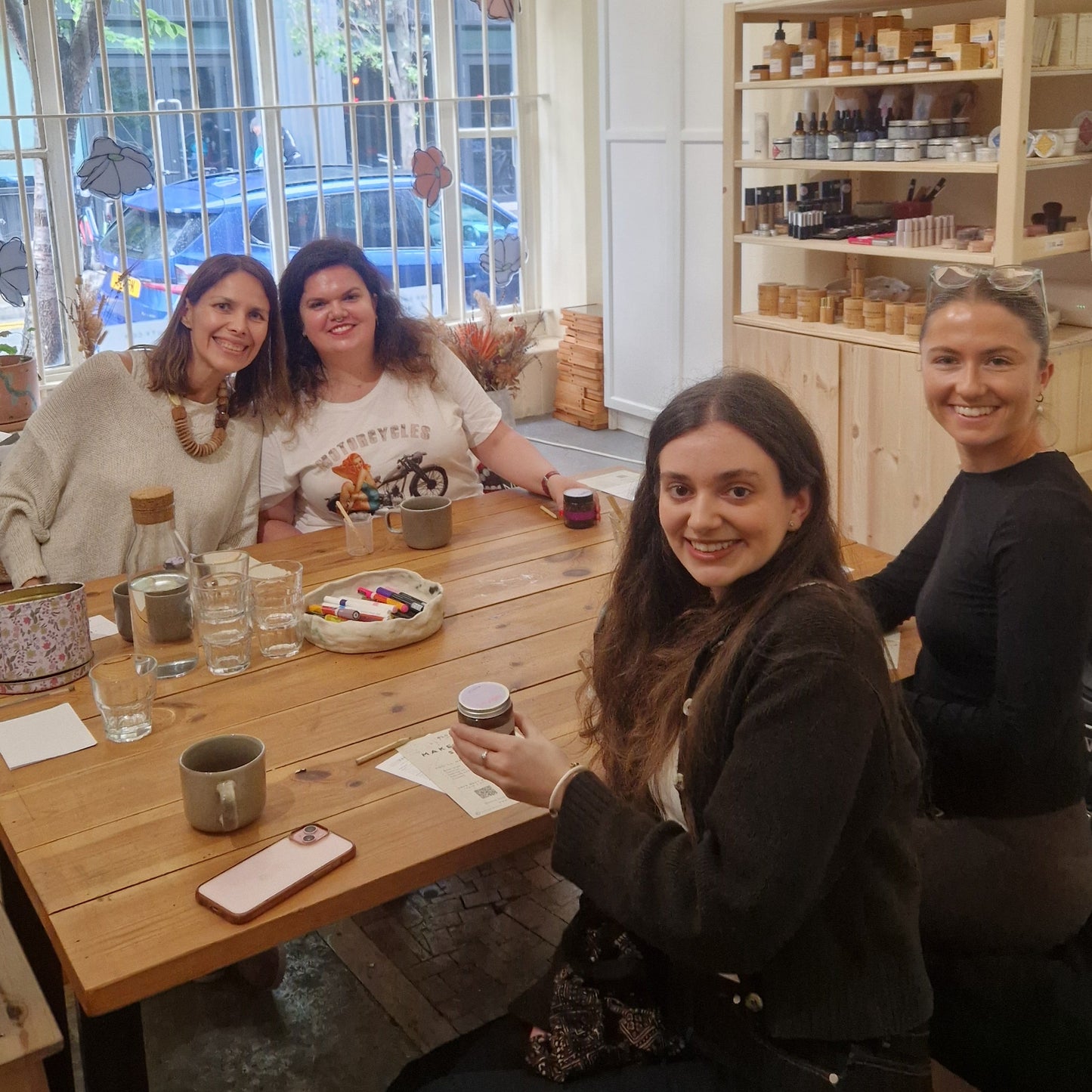group of women sitting at a table smiling and holding up the body scrubs they have made and decorated as part of a private birthday celebration activity in london