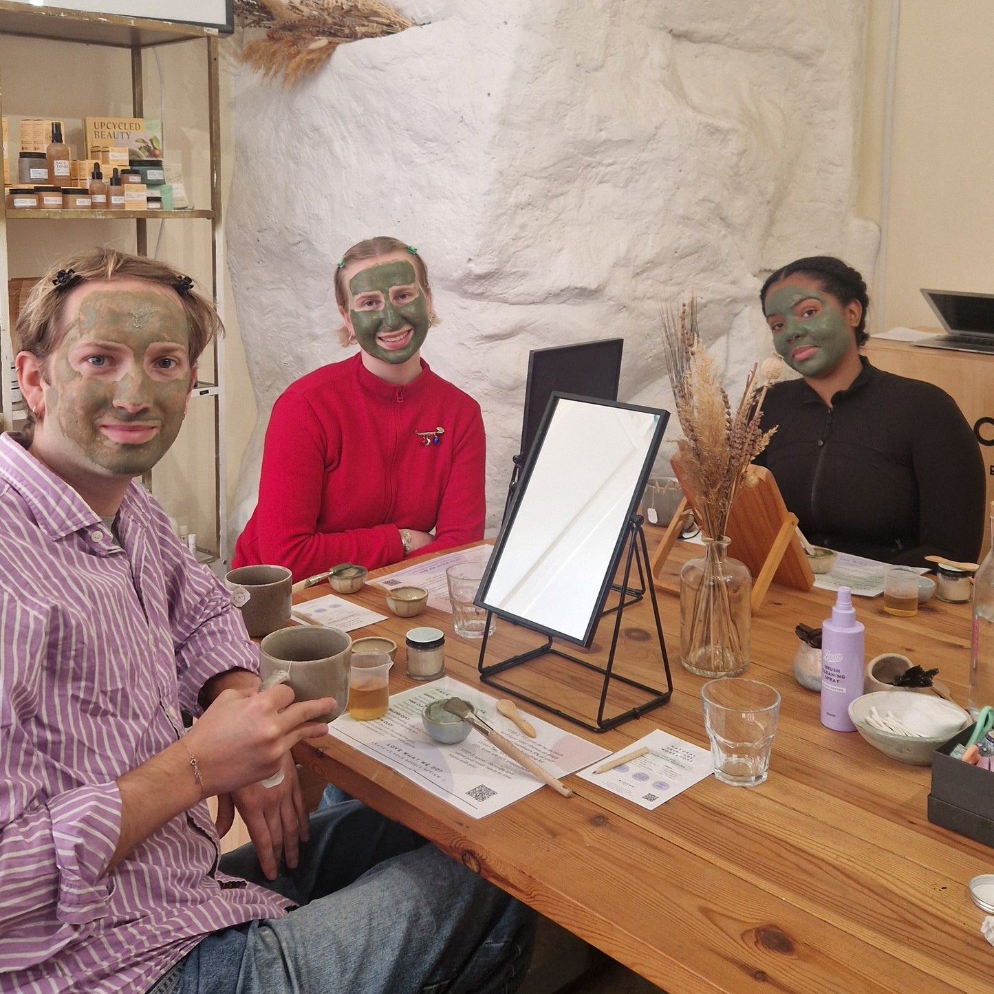 three people with clay face masks on sitting around a table with mirrors in blomma beauty's natural skincare studio in london