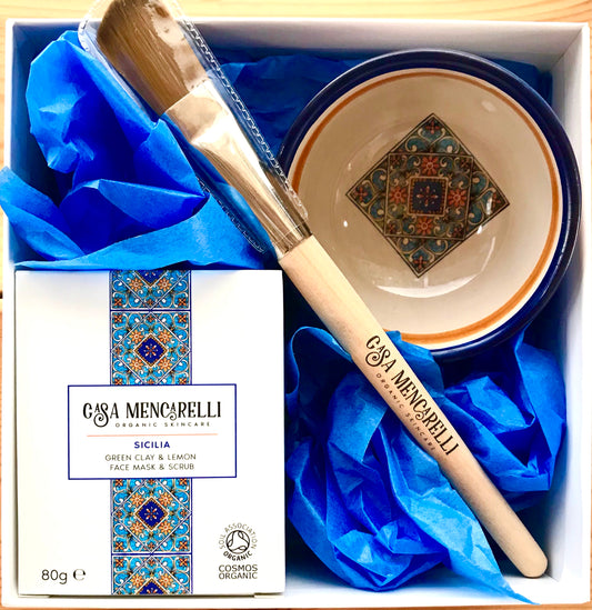 a white square box housing a hand painted umbrian ceramic bowl which is a creamy colour and with orange and blue accents. a wooden face mask applicator brush and a white box with blue and orange tile design which contains a green clay and powdered lemon peel face mask