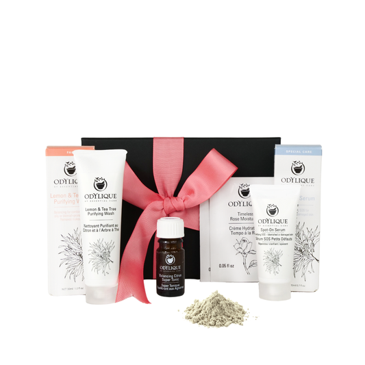 photo of natural skincare set on a white background. The skincare set is from organic skincare brand odylique. the kit comes in a black kraft box with a pink ribbon which is in the background. the organic skincare products are in the foreground and includes a lemon and tea tree face wash, a citrus tonic, two sachets of rose moisturiser and a white tube of spot on serum for blemishes. there is a small pile of grey/green powder to show the texture of the clay and maca face mask