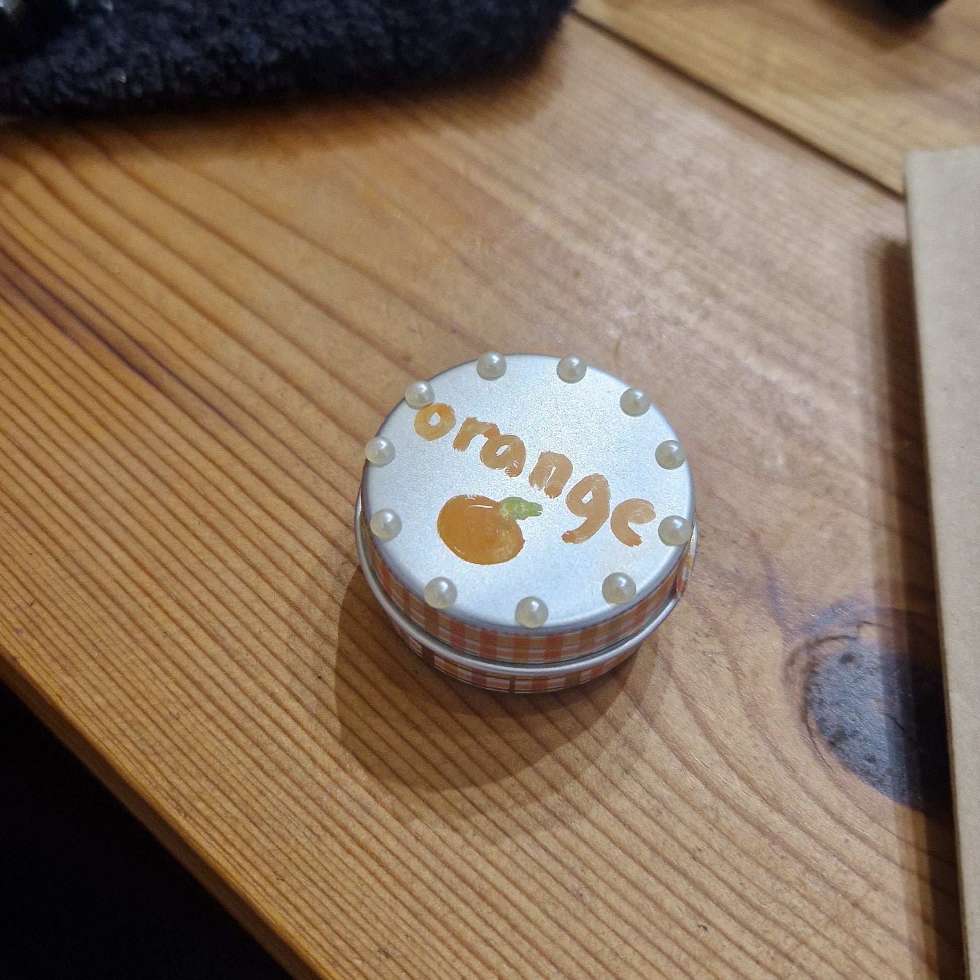 Small round container labeled 'orange' on a wooden surface with pens and a cloth.