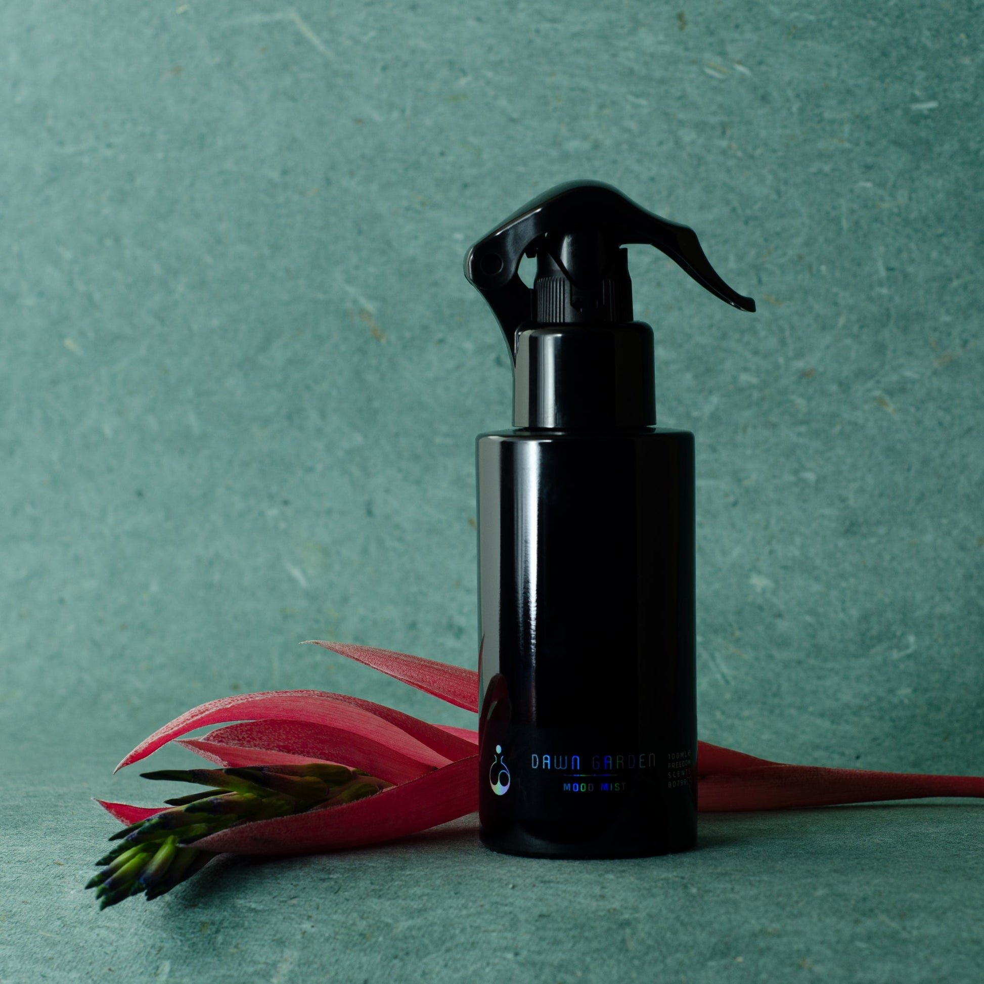 home fragrance in a black spray bottle on a teal background with tropical flower next to the bottle. freedom scents hand made natural home mists