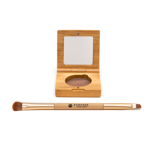 photo of bomonde mineral make up's eyebrow palette and double ended eye brush on a white background. the eyebrow powder is pressed into a round pan and encased in a bamboo compact case with mirror. the brow powder is dark brown. in the foreground there is a double ended eye brush made from gold metal and bamboo. the brushes are synthetic bristles. one end is a fluffy eyeshadow brush and the other end is a compact, angled brush for eyeliner and eyebrow powder