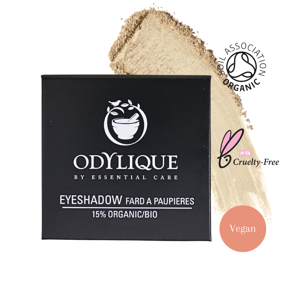 A black square box of Odylique Eyeshadow with white text, shown next to a beige powder smear on a white background. The packaging reads 'ODYLIQUE BY ESSENTIAL CARE EYESHADOW FARD A PAUPIERES 15% ORGANIC/BIO' and includes certification and vegan badges.