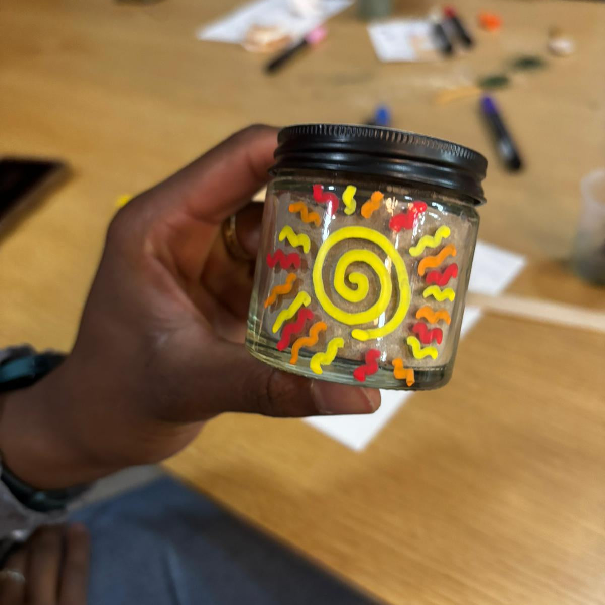 Hand holding a jar with colorful spiral design on a wooden table.