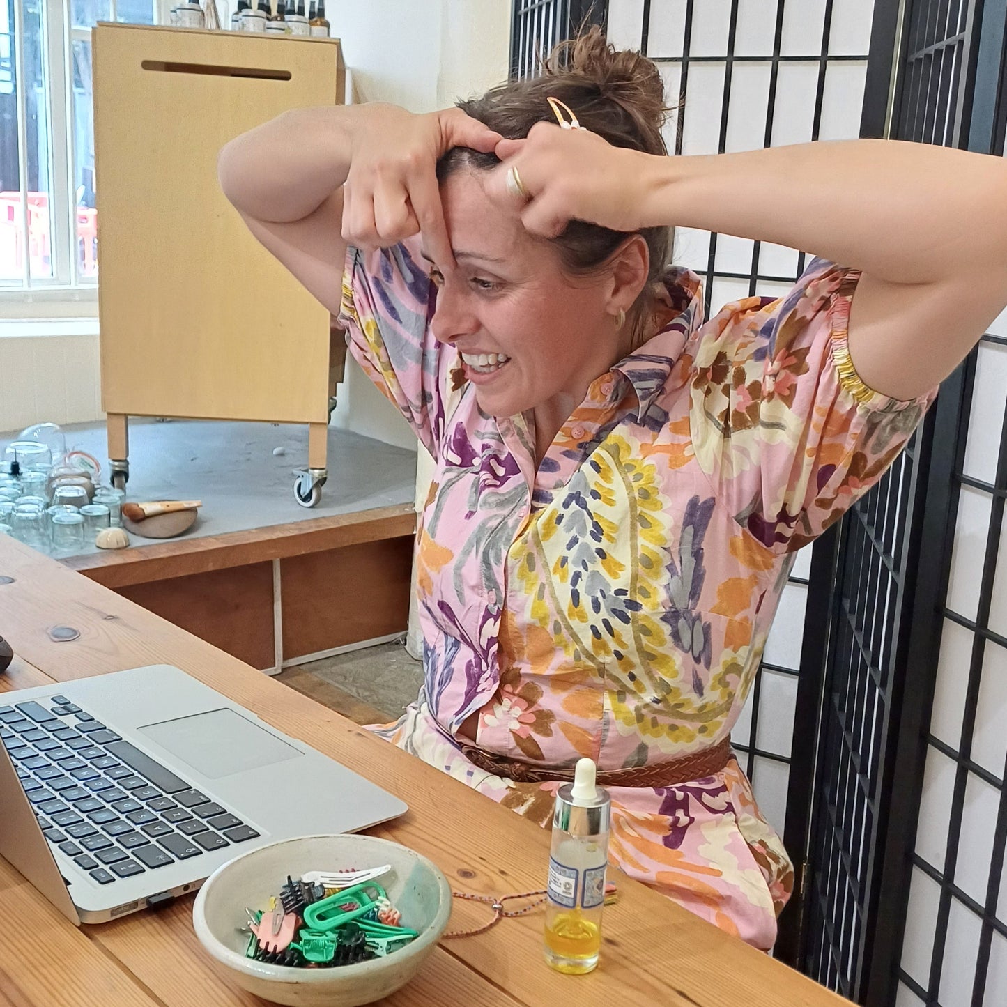 Person sitting at a desk with a laptop, phone, and water bottle in a room with a window as she demonstrates face massage techniques to online students