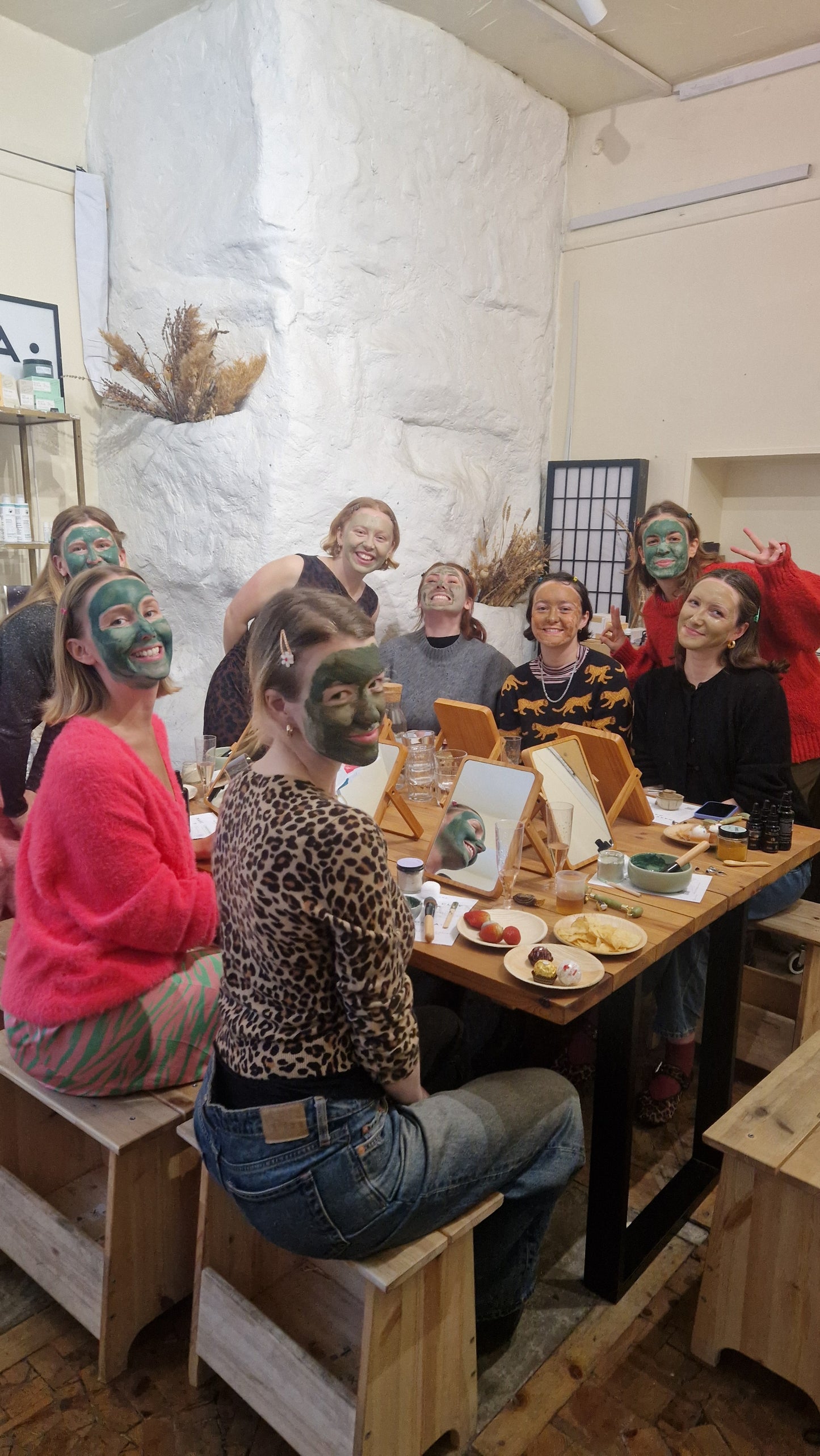 group of women with clay face masks on sitting around a table with mirrors, skincare products and snacks as part of a birthday party, hen party or baby shower celebration