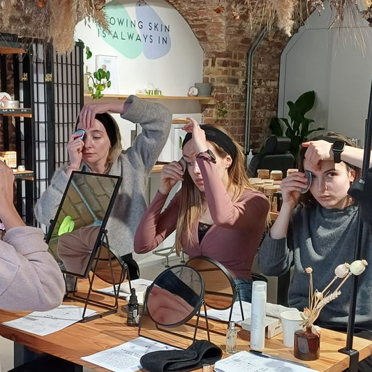 3 people sitting with headbands in front of individual mirrors using gua sha stones to learn how to do a gua sha facial