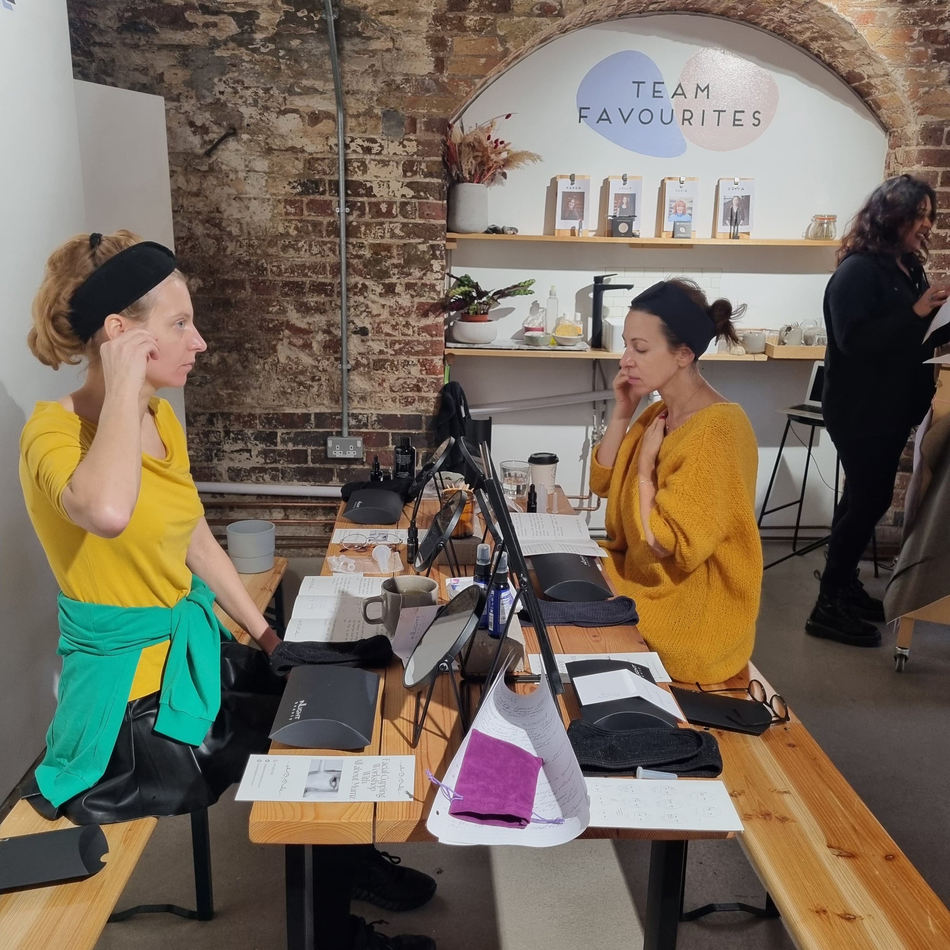 two women sitting opposite each other both with yellow tops and head bands on. they are practising using facial cups at the natural skincare workshops at blomma beauty