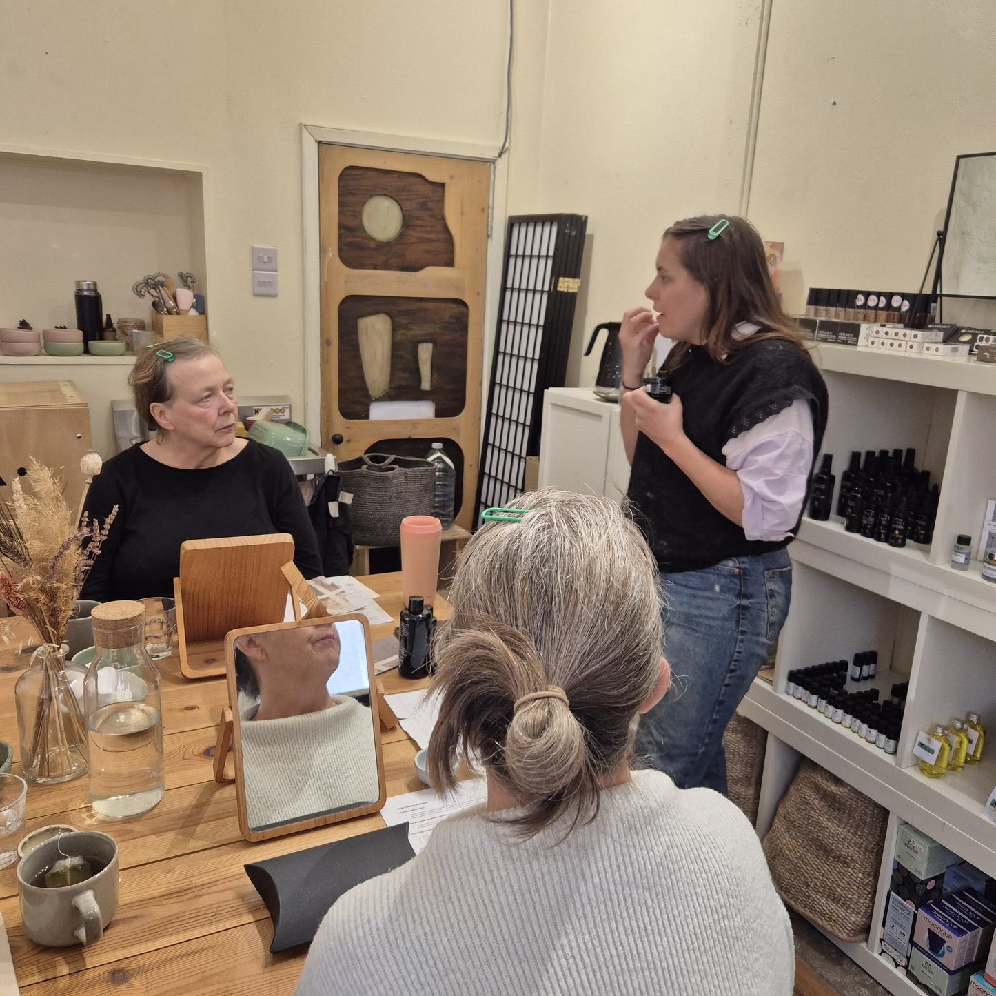 people sitting at a wooden table surrounded by mirrors and beauty products listening and watching a teacher demonstrating natural skincare techniques