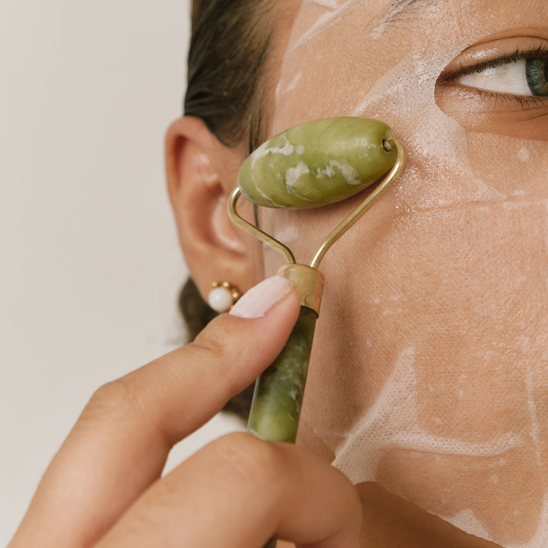 woman with brown hair can be seen using the jade facial roller on top of a white sheet mask