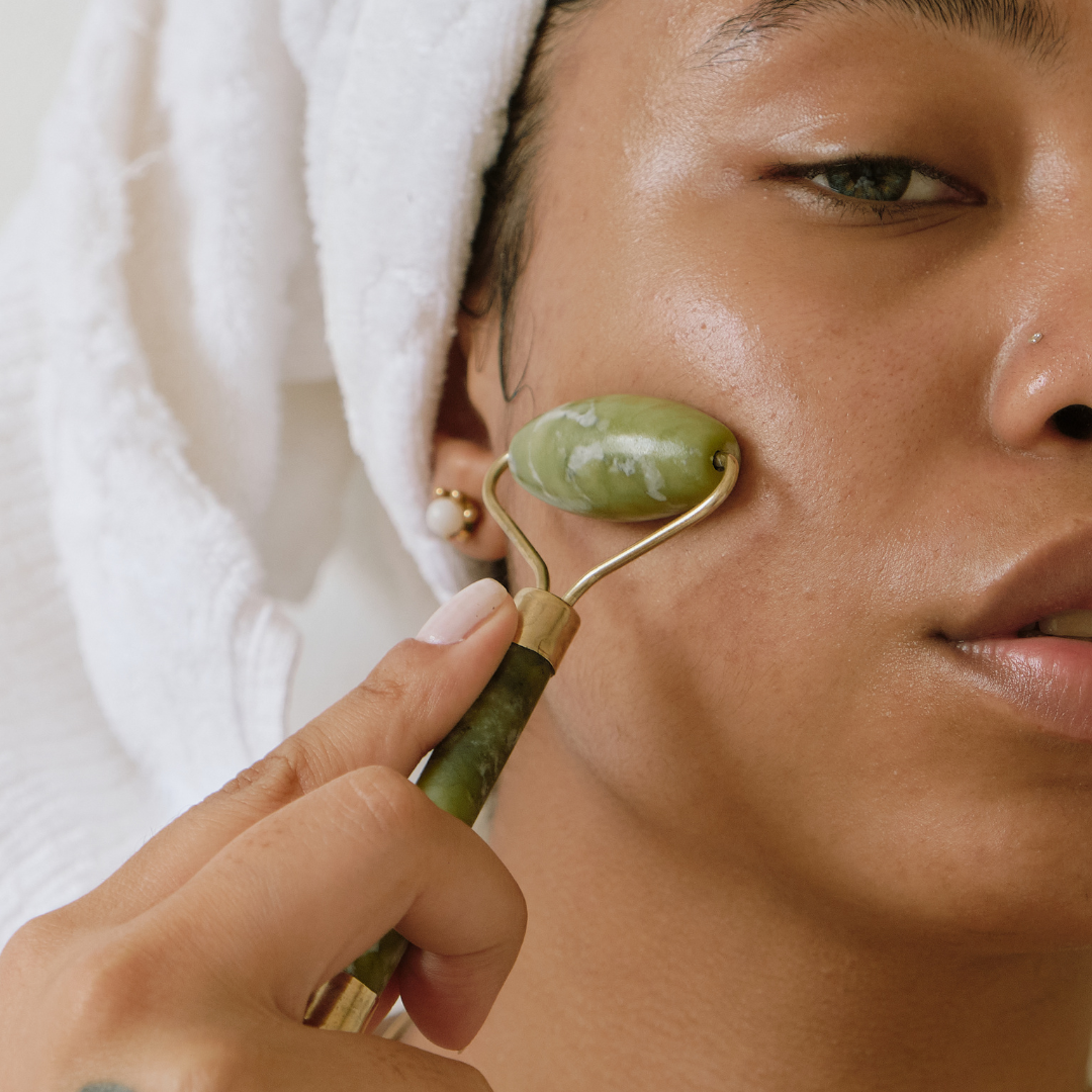 woman with brown skincare and hair wearing a white towel on her hair can be seen using the jade face roller to massage her face