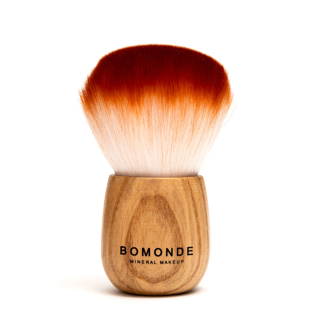 photo of bomonde mineral make up's kabuki brush on a white background. the brush has fluffy bristles that are more orange at the top and creamier as they get to the wooden handle which is made of bamboo and has the bomonde logo printed on it in black
