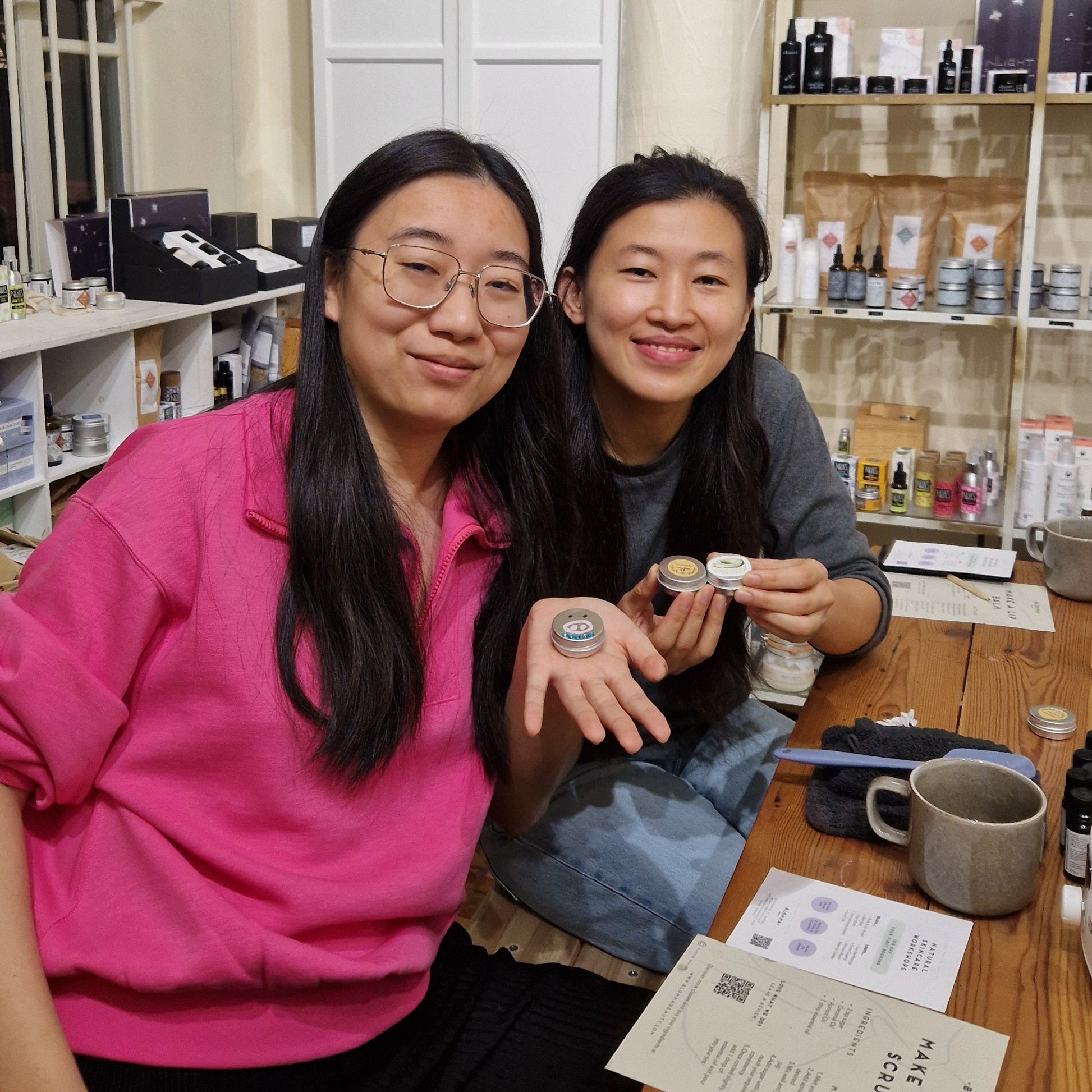 Two women sitting at a table in a store, smiling and holding their lip balms which they have made and decorated as part of an experience at blomma beauty's natural skincare studio