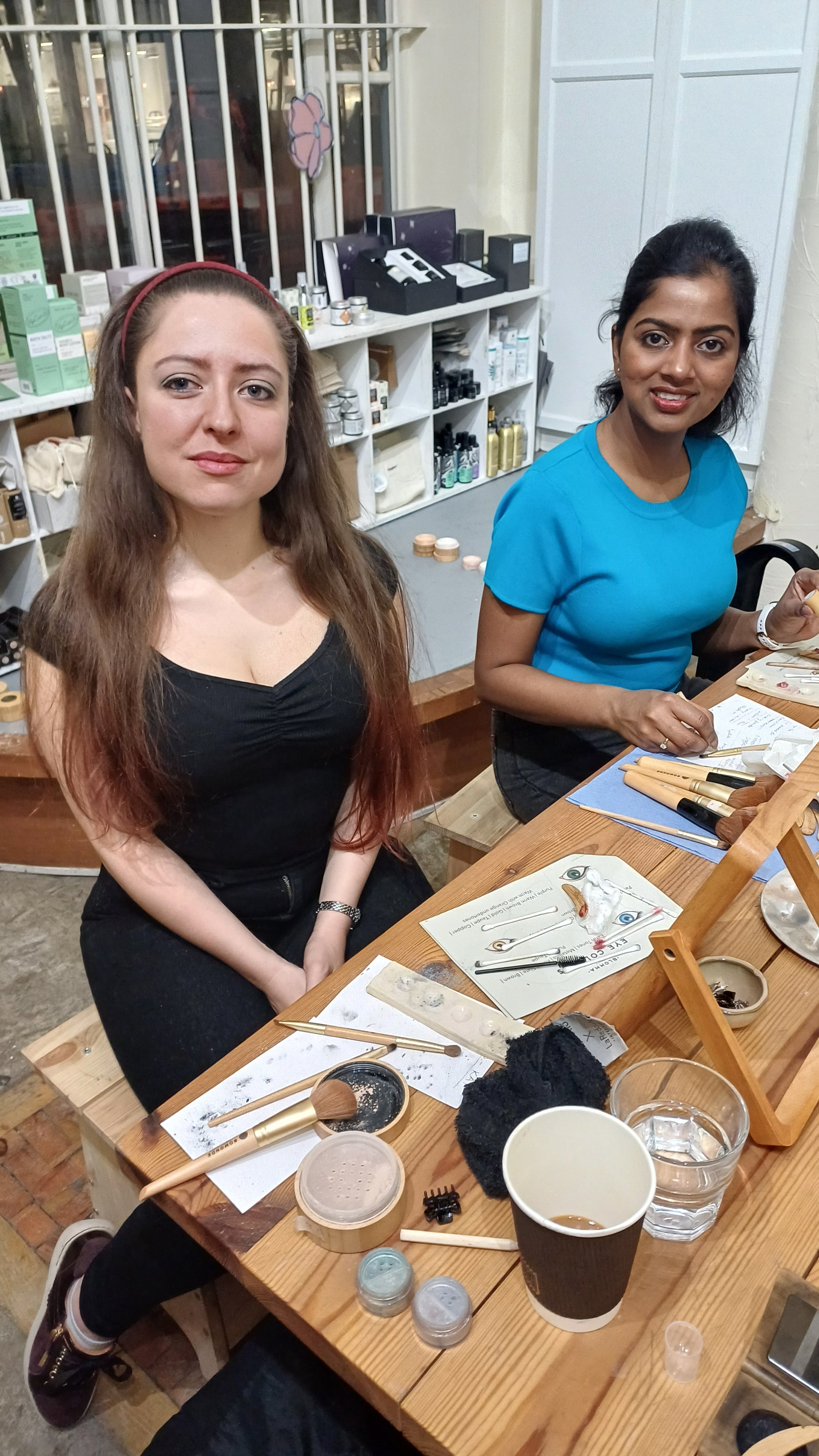Two women standing at a table with various items on it in blomma beauty's natural makeup studio in london. they are attending a makeup masterclass to learn makeup application techniques