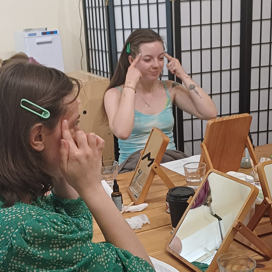 two women sitting in front of mirrors in the blomma beauty skincare studio in london taking part in a face massage class. they are holding their hands up to massage their temples