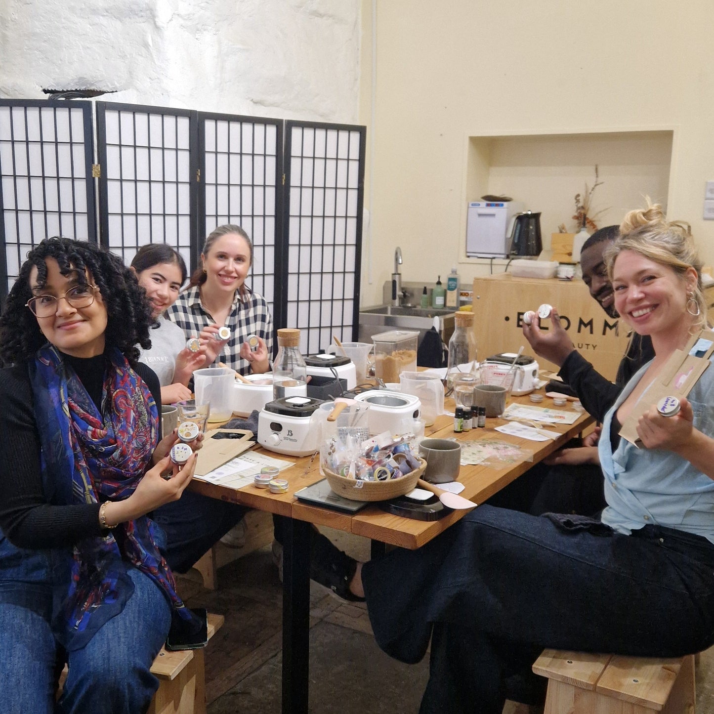 group of people sitting around a wooden table in a small studio in london where they are holding up the lip balms that they have made and decorated