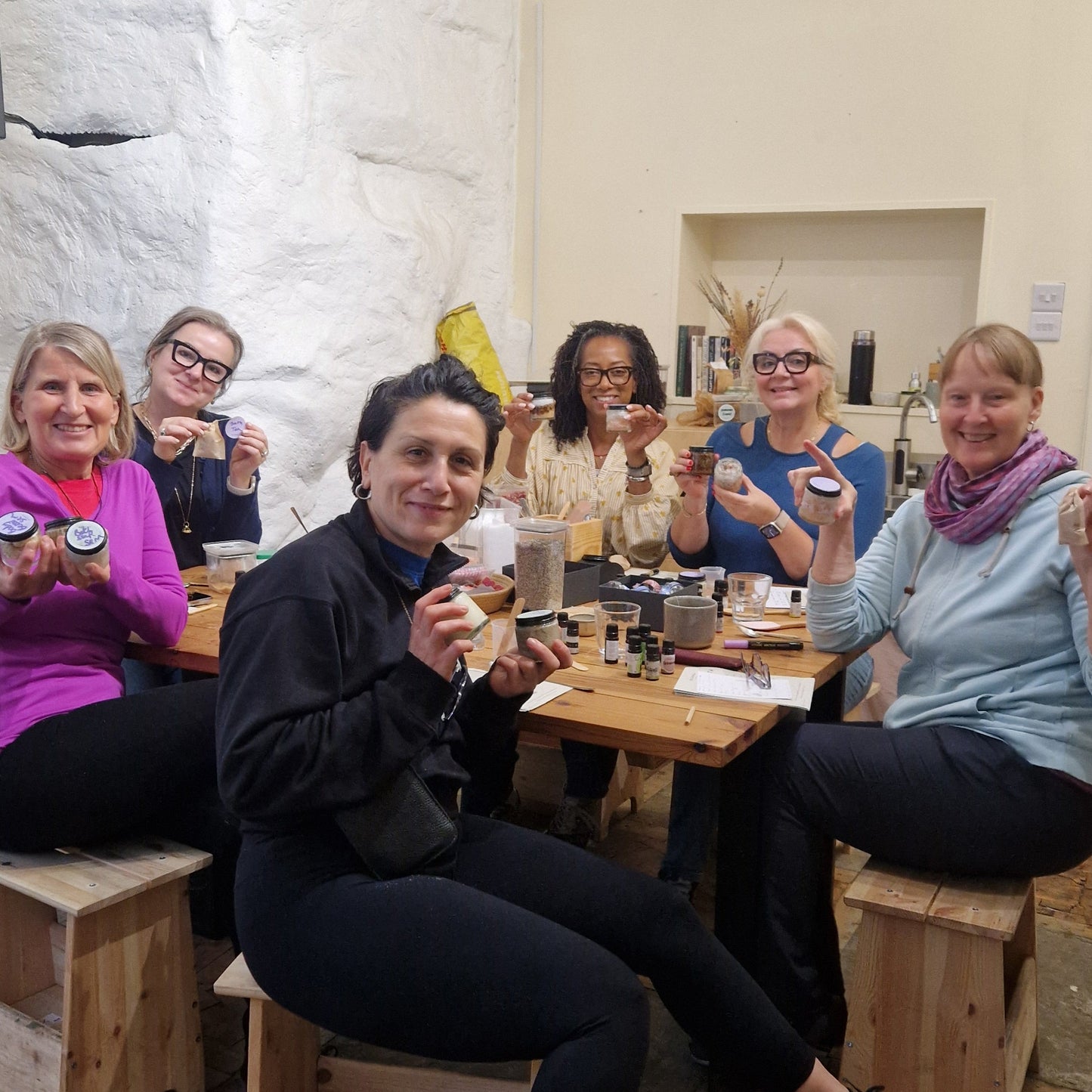 group of people sitting around a table showing the products they made at a skincare making class 