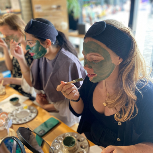 Three individuals sit at a table. They are wearing black headbands and are applying green facial masks. The table has small containers of facial products and mirrors. They appear focused on applying the masks evenly to their faces.