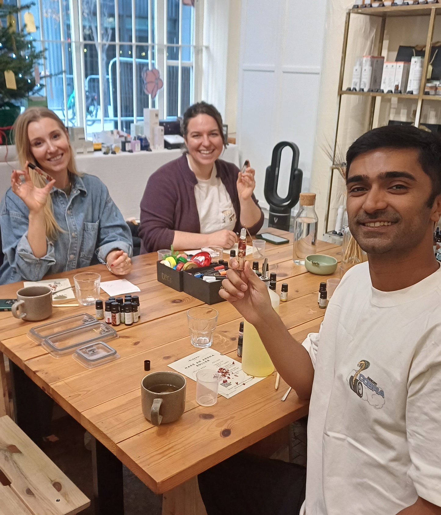 group of people sitting around a wooden table showcasing their perfume rollerballs made at a blomma beauty workshop in london