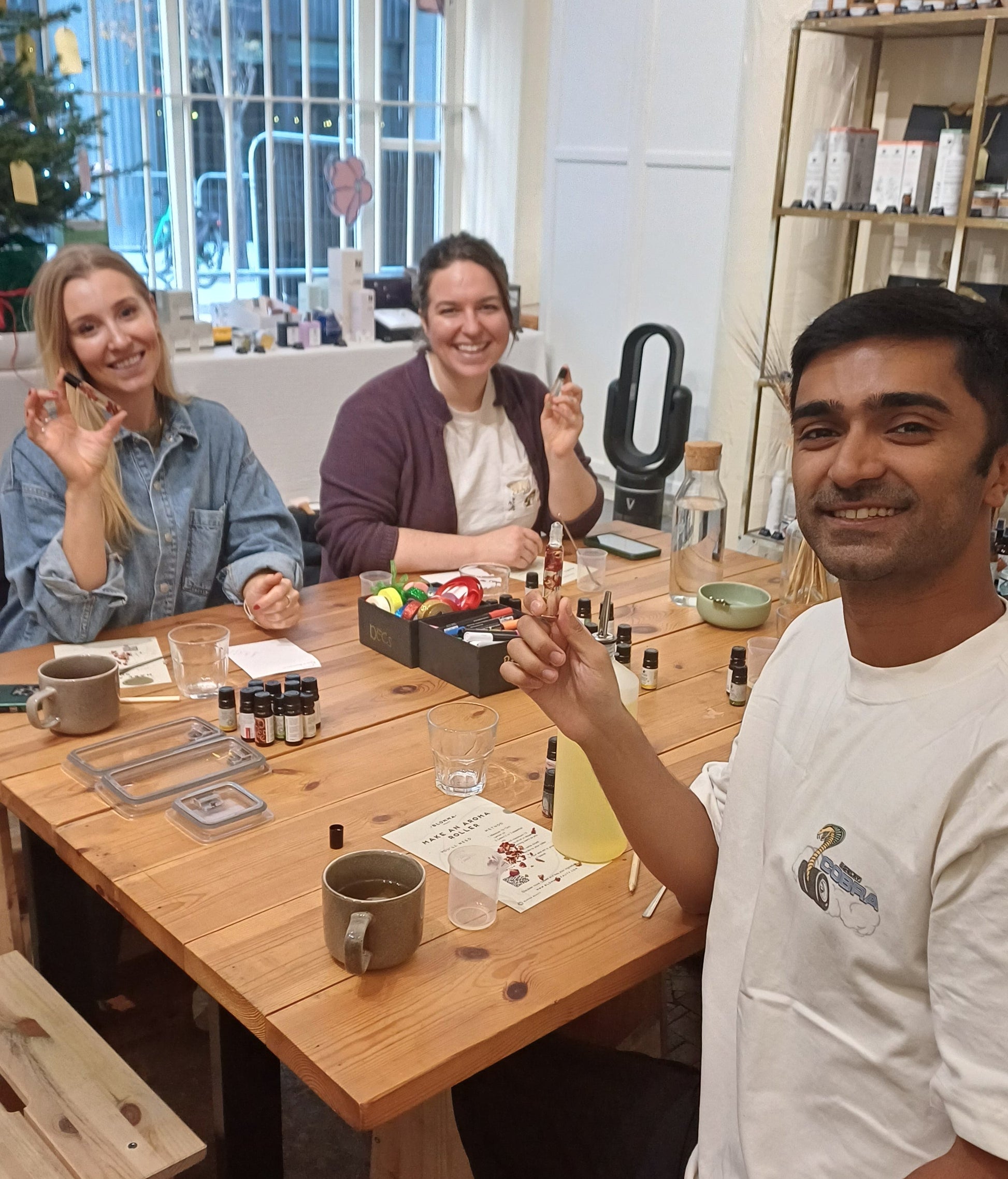 group of people sitting around a wooden table showcasing their perfume rollerballs made at a blomma beauty workshop in london