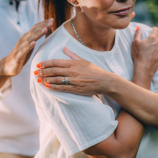 woman sitting with arms crossed across her chest with a person holding hands on her shoulders from behind conducting reiki energy transfer on her