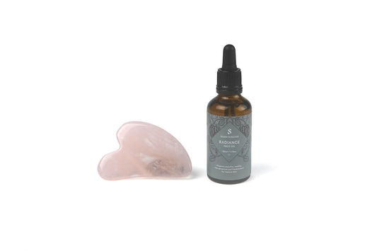 a pink rose quartz gua sha facial massage tool is displayed next to silvan skincare's radiance face oil which is in a brown amber pipette bottle with blue-grey label on a white background