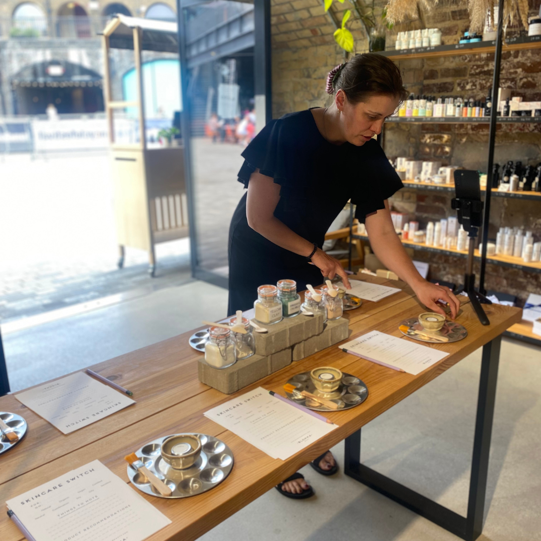 Blomma owner, Karen, is shown in a black dress, setting up a workshop table on the Blomma shop floor. The table is filled with pots and jars for creating/testing facial masks.