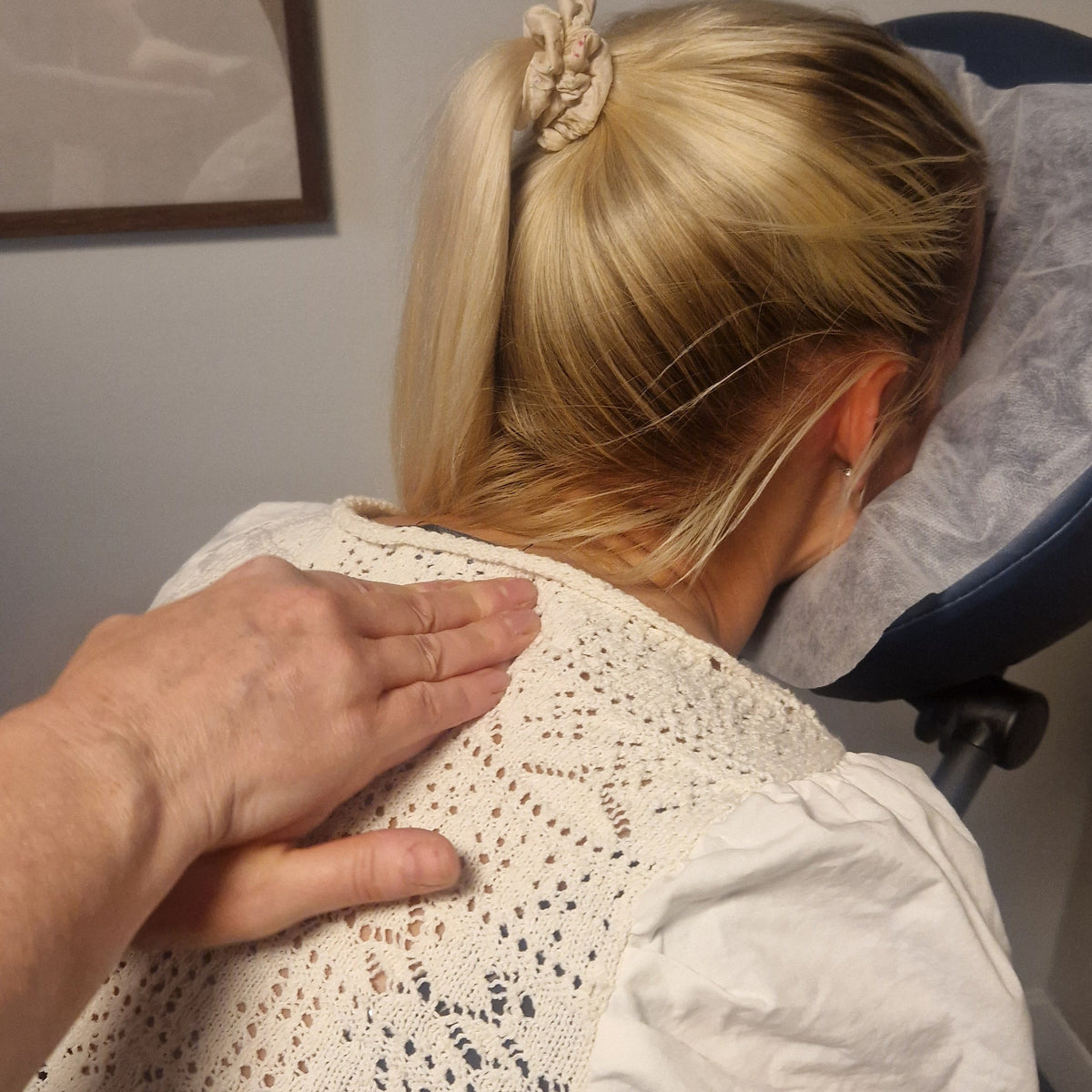 A woman with blonde hair in a ponytail leans forward into a massage chair while another person's hand rests on her upper back. She is wearing a white short-sleeve top with a textured crochet pattern.