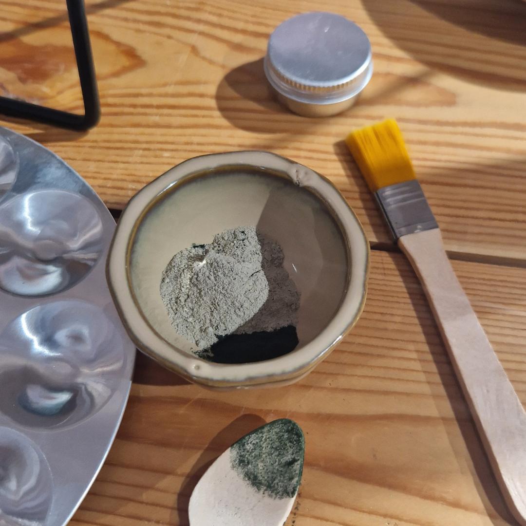 A small pot on a wooden table is filled with a powder face mask. Around the pot is a small face brush, a small aluminium pot and a wooden spoon, the spoon is stained with green powder.
