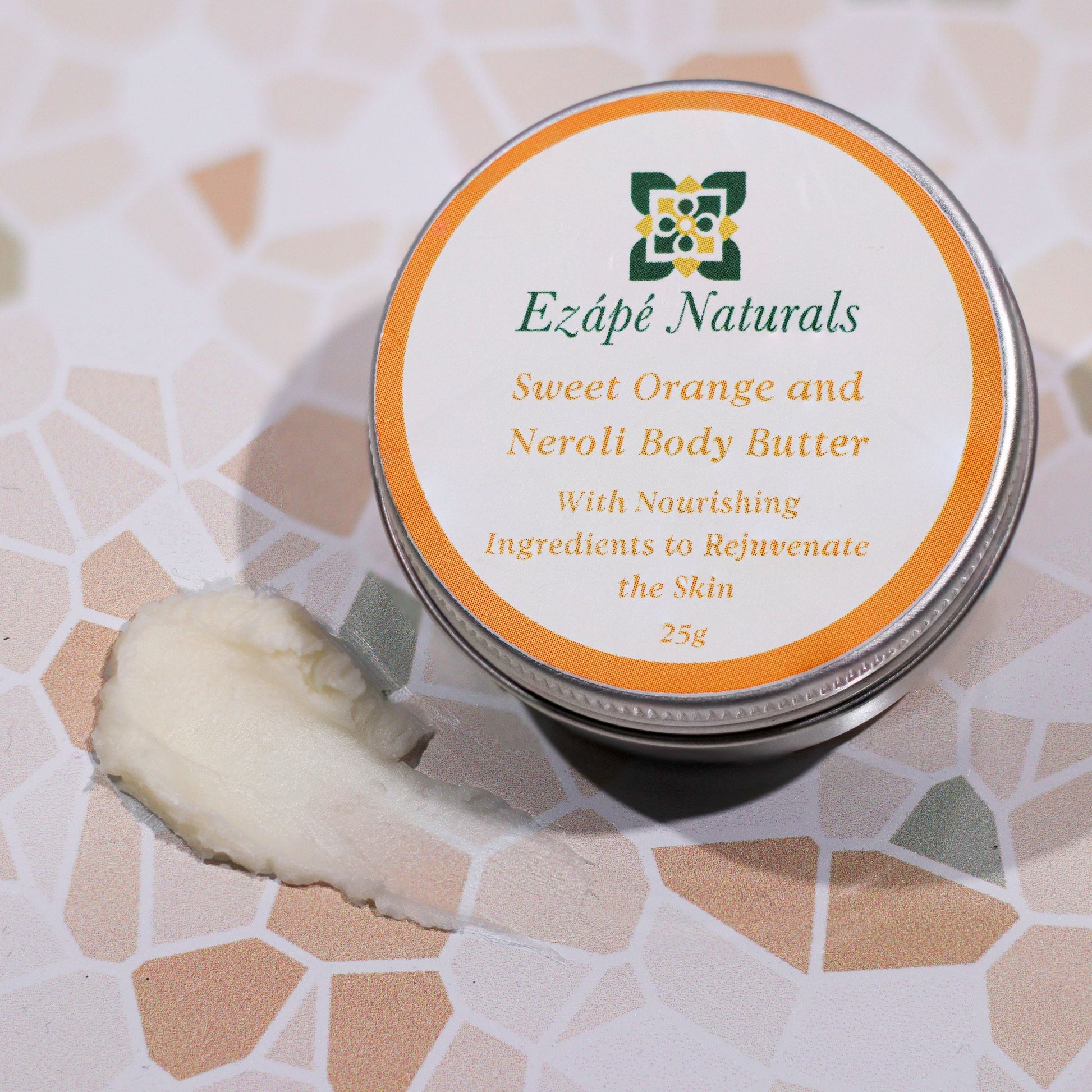 Ezape Naturals Sweet Orange and Neroli Body Butter on a textured surface that looks like a mosaic. there is a slick of body butter showing so you can see the texture and then then small round aluminium tin that it comes in which is labelled with a round white sticker with an orange border