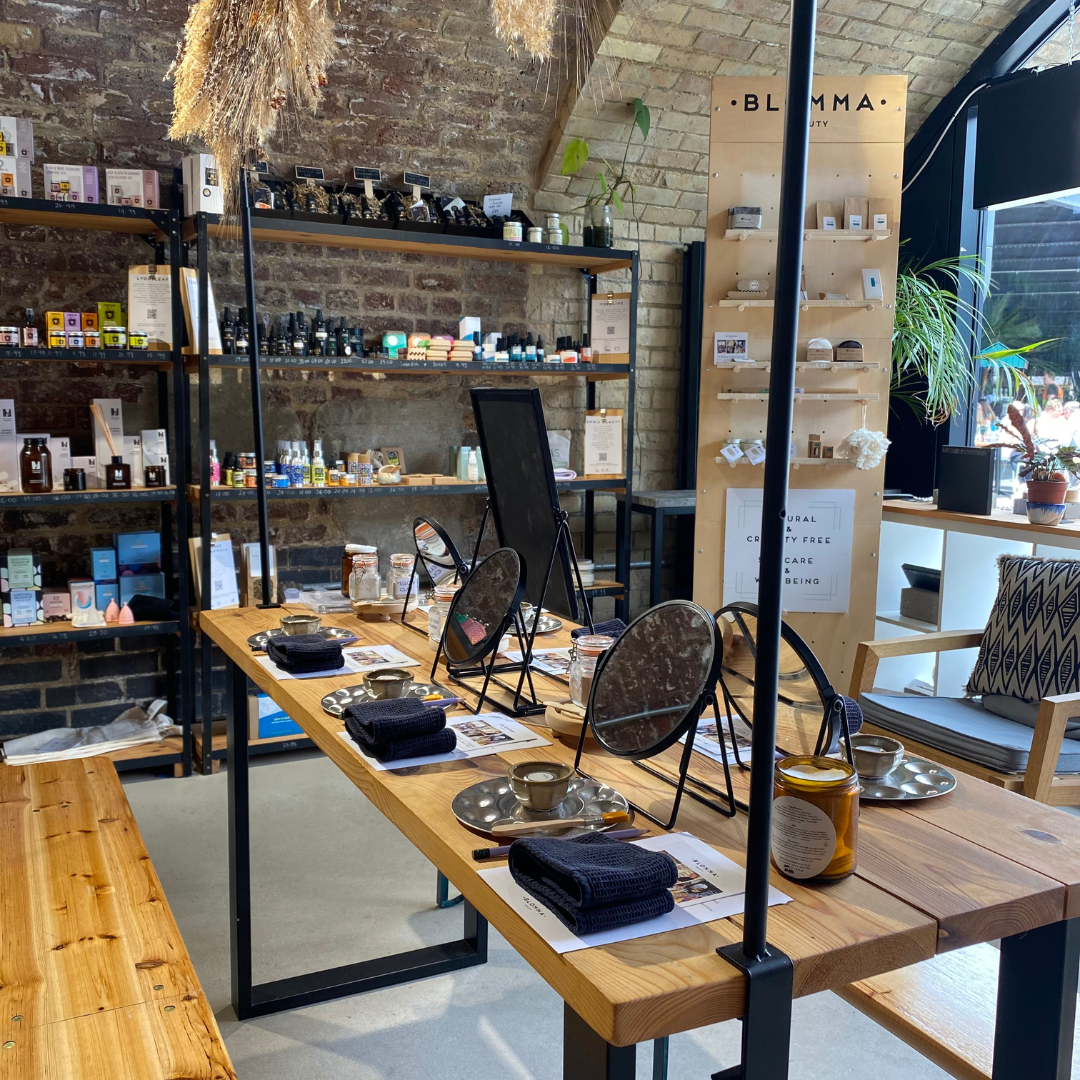 Cozy, well-lit interior of the Blomma store. The wooden table is set for a workshop, with round mirrors, tin trays, towels, and various small containers. The shelves lined with clean beauty products and the brick wall enhance the warm and inviting atmosphere.