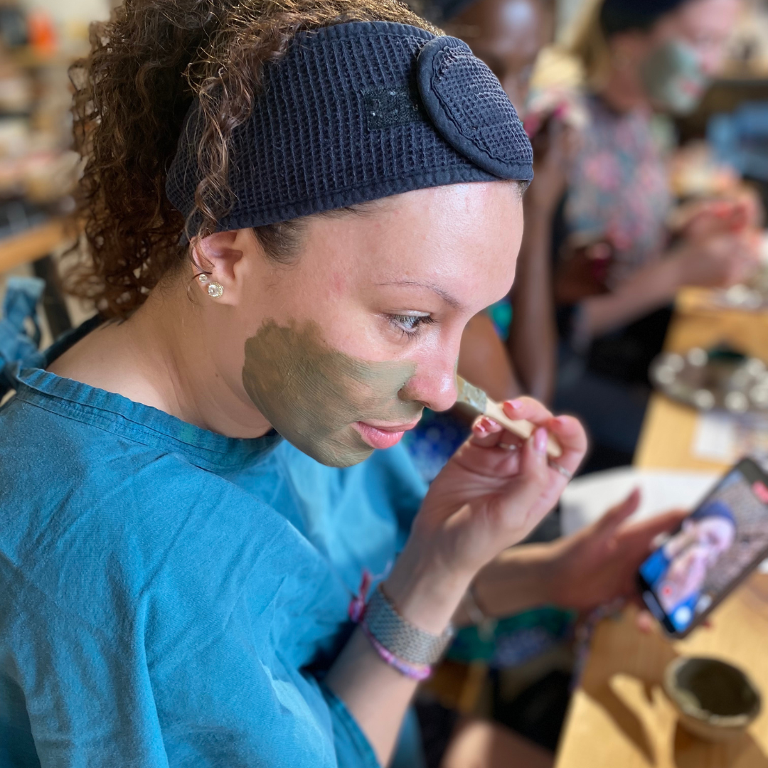 A person is holding a small brush and using it to apply a green face mask to their cheek. They are wearing a black headband and a blue gown. There are other people in the background also engaged in the same activity.